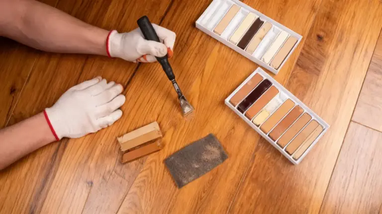 Mani con guanti che riparano un graffio su parquet in legno con attrezzi e cere colorate